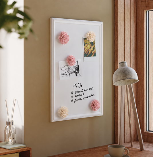 A white rectangular magnetic cork board with a white frame hangs on a warm beige wall in a styled room. The board is decorated with pink and cream pom-poms and pinned with a small art print, a sketch, and a handwritten to-do list. - alternate view